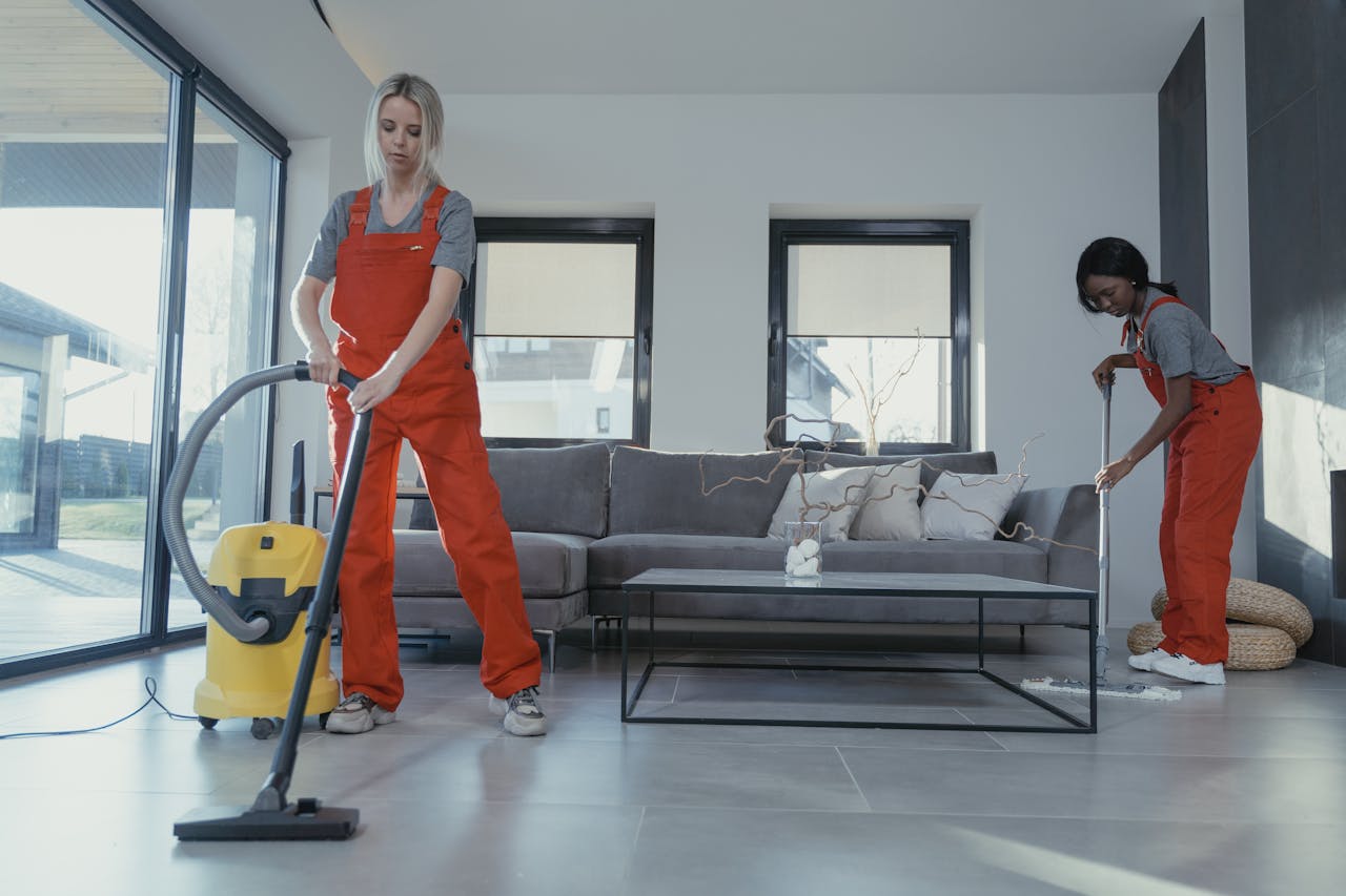 heros-img Two women in red overalls vacuuming and sweeping a modern living room.