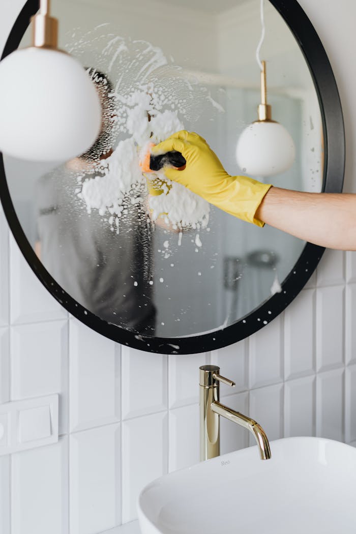 about-us-02 A person wearing a yellow glove cleans a bathroom mirror with foam spray.
